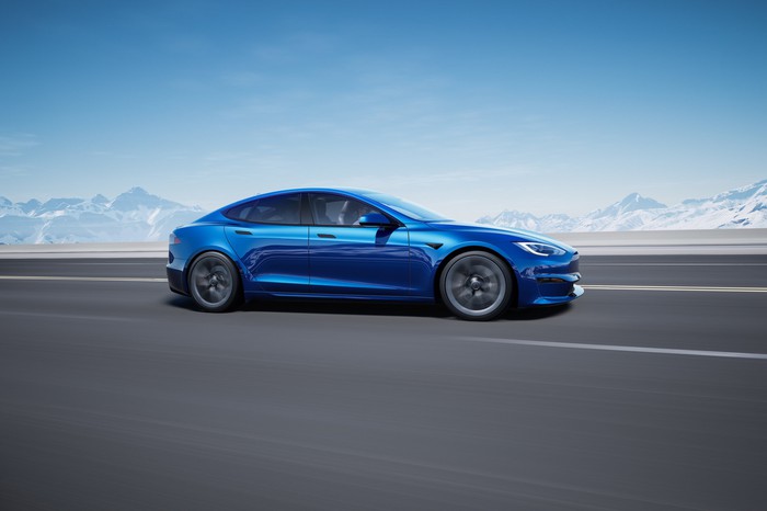 A blue Tesla Model S cruises down an open road with mountains in the background.