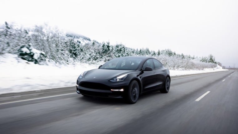 A Tesla Model 3 drives on a snowy road. We see it from a front three-quarter angle. The car is sleek, in glossy black with black wheels and trim.